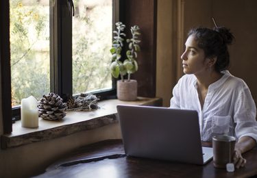Mixed-race young adult woman sits at a table, using her laptop computer, with a cup of tea also on table. She is deep in thought, gazing out the window. A houseplant, candle, and pine cones are on the windowsill. Filtered sunlight reveals yellow flowers outside; natural light also illuminates woman's profile. Woman has dark brown hair twirled into a bun. She's wearing a white shirt with sleeves rolled up. Working from home on a deadline, on track to get the project done on time and under budget. Creative problem-solving is her specialty and she does it best from her home office.