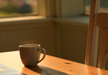 A hot cup of coffee in the veranda at a beach house. Coffee is backlit, hot, and steaming as it sits on a rustic wood table in a cabin. Themes include coffee, caffeine, cup, beverages, hot drinks, morning, relaxing, ceramic, porch, rustic, country, hot chocolate, espresso, Americano, latte, cappuccino, and steam.